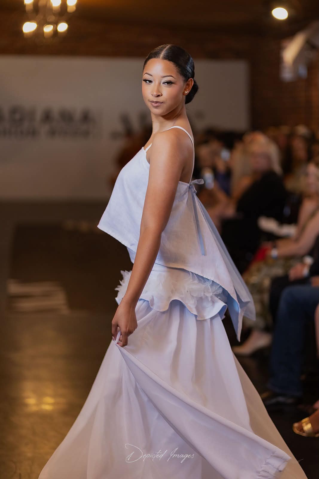 A model walks the runway wearing a white linen and chiffon top and skirt set with a layered, ruffled design and a draped style back to the top, showing the light blue underlining. The background shows a blurred fashion show venue with an audience and a runway.