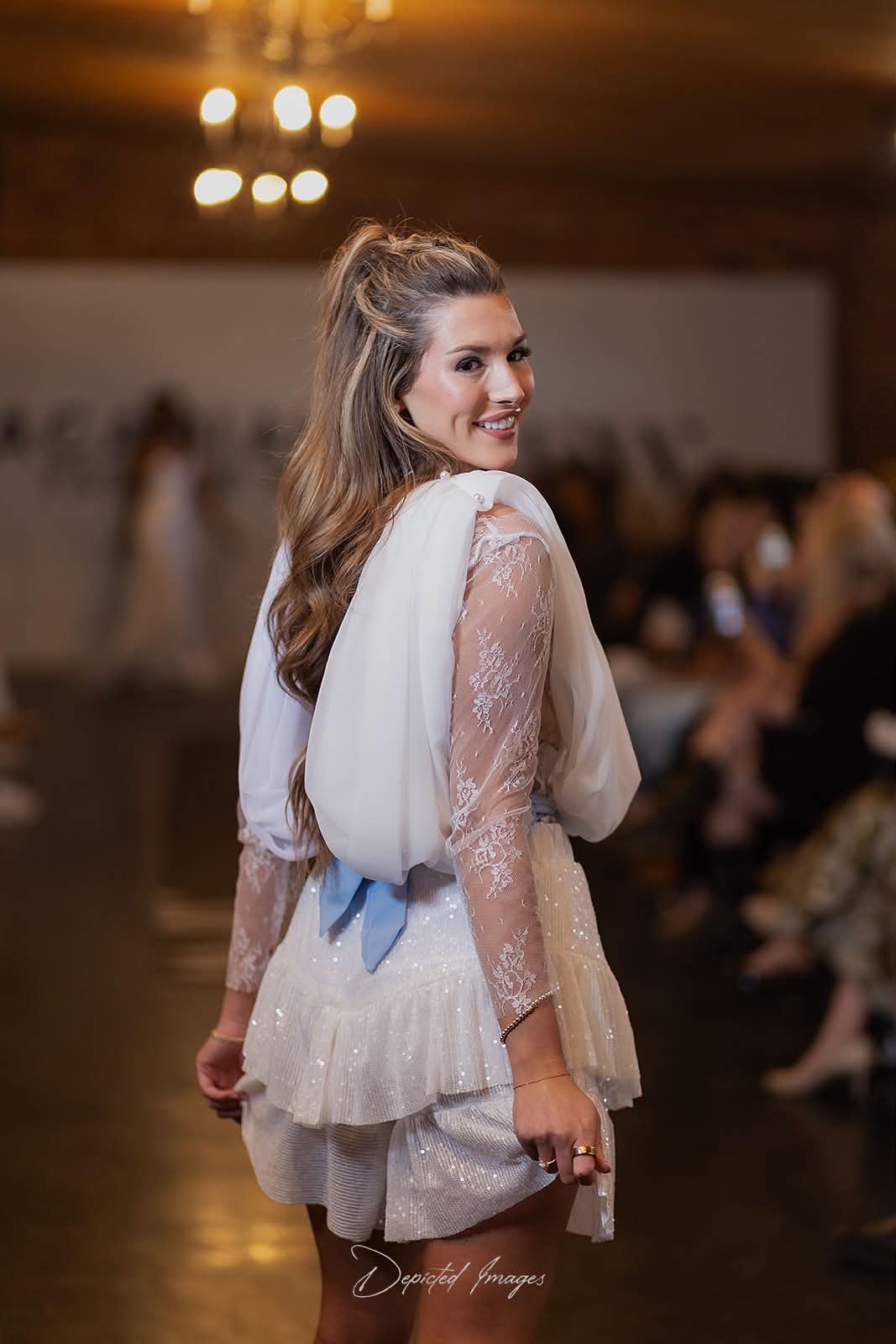 A woman on a runway wearing a white lace long sleeve undershirt with sheer lace long sleeves, a ruffled hem short, and a light blue bow at the back of the white chiffon draped over-top, photographed in a fashion show setting.