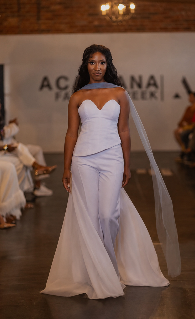A model walks down a fashion runway wearing a strapless white dress with a structured bodice, wide legs, and a long sheer veil. The runway is indoors with seated spectators on both sides and a branded backdrop behind the model.