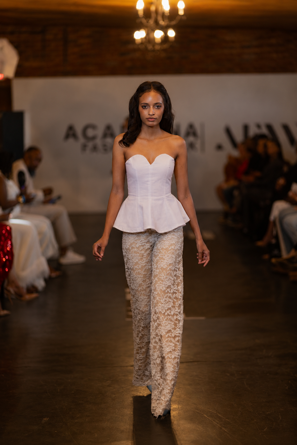 A model walks the runway wearing a white strapless sweetheart corset top paired with matching cream lace wide-leg pants. The runway show is held in a venue with a wooden floor, brick walls, and an audience seated on both sides.
