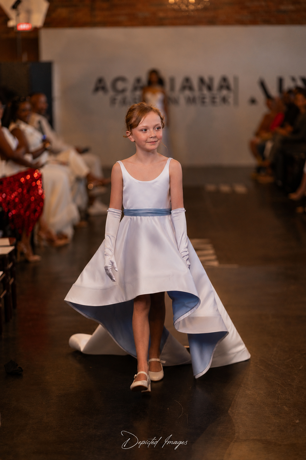 A young girl walks down the runway wearing a white sleeveless dress with a light blue sash and matching long gloves in a fashion show setting.