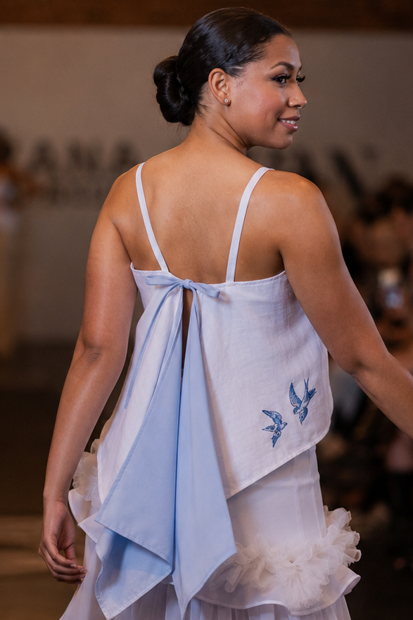A woman on a runway wearing a light blue and white sleeveless dress with a large draped back detail and ruffled layers, photographed in an indoor fashion show setting.